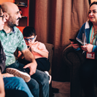 photo of casually dressed in-home care provider wearing a name tag and holding a notebook and pen speaks to a couple and their two children